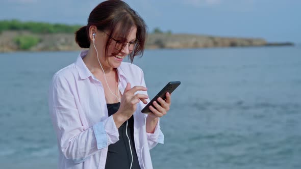 Happy Middle Age Woman Relaxing on Beach Wearing Headphones with Smartphone alt