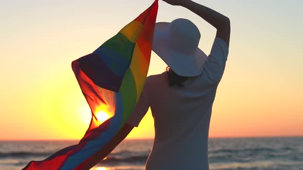 Back View of Woman in White Dress and Hat Holding Gay Pride Flag at Sunset in Beach alt