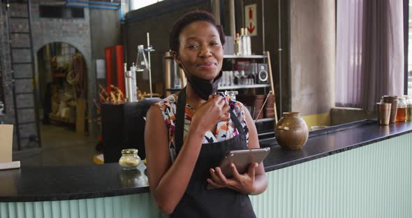Portrait of african american woman in face mask working at a bar, using tablet and smiling to camera alt