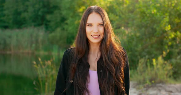 Portrait of a Smiling Young Woman with Long Dark Hair Outdoors in a Forest Near a Lake alt