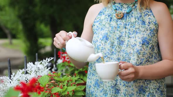 Woman in a Summer Cafe Pours Tea Into a Cup Close-up Standing. Woman Stands on the Background of the alt