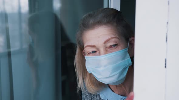 Portrait of Lonely Elderly Woman in Quarantine with Face Mask Looking Through the Window alt