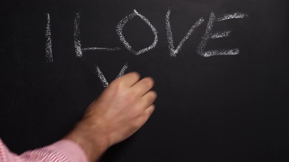 A man's hand in a shirt writes in white chalk on a black board I love you alt