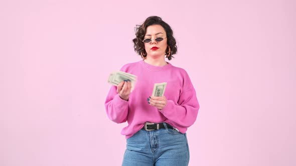 Stylish avarice woman counts a wad dollar money isolated on pink background. alt