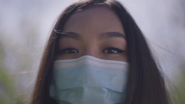 Headshot of Asian Woman in Covid19 Face Mask Looking at Camera Standing Outdoors in Park alt