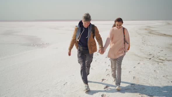 Happy Lovely Couple Travelers Walking Hand to Hand on Pink White Salty Coast alt