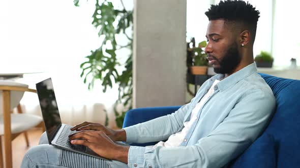 Smiling AfricanAmerican Freelancer Guy is Using Laptop Computer Sitting on the Couch at Home alt