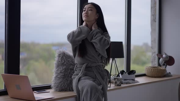 Young Asian Woman Stretching Neck Sitting on Windowsill in Home Office alt