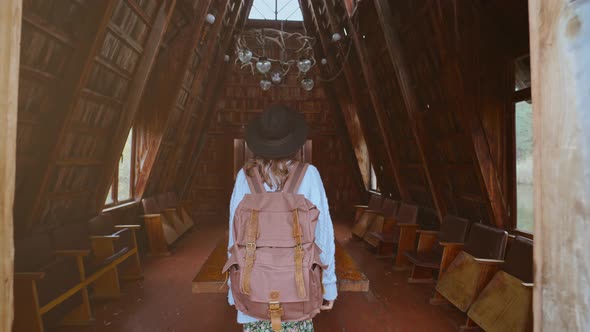 Stylish Tourist Woman in Hat with Backpack Coming in Old Vintage Style Wooden Hut alt