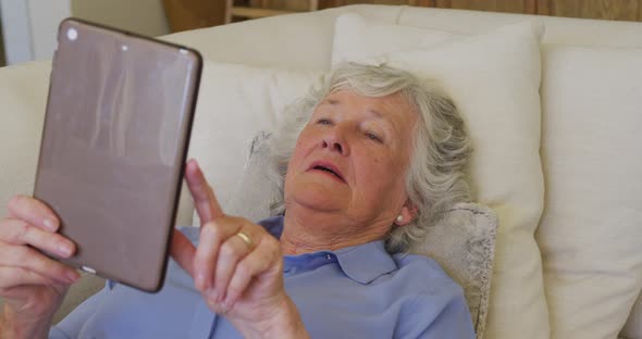 Caucasian senior woman smiling while using digital tablet lying on the couch at home alt