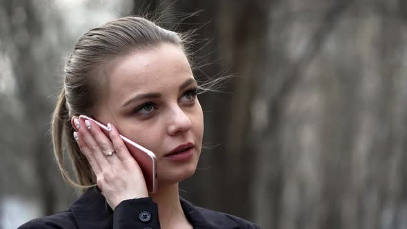 Young White Girl in a Black Jacket Talking on the Phone in a City Park alt