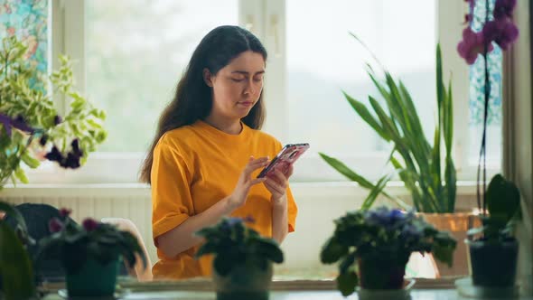Woman chatting on the smartphone sitting near the window at home. Interior with natural plants alt