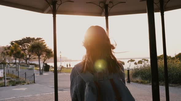 Camera Follows Happy Casual Young Woman with Backpack, Hair Blowing in the Wind Walking Along Summer alt