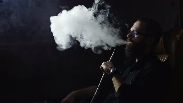 Young bearded man smokes a hookah and blow out smoke on black background in slowmotion