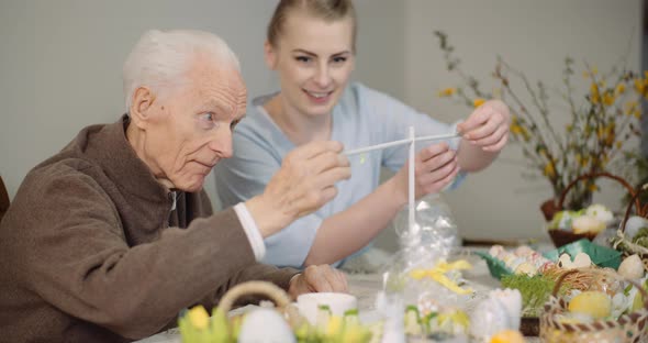 Senior Man and Granddaughter Decorating Table With Easter Eggs and Easter Decorations. alt