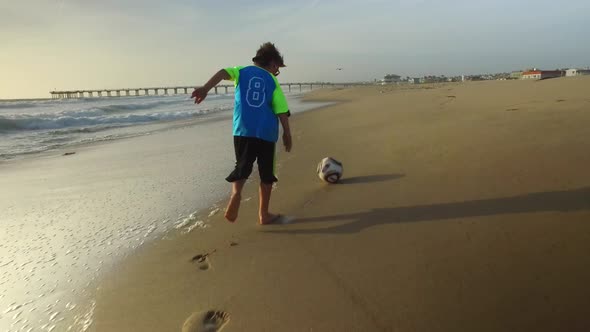 A boy kicks a soccer ball on the beach at sunset with the ocean and pier. alt
