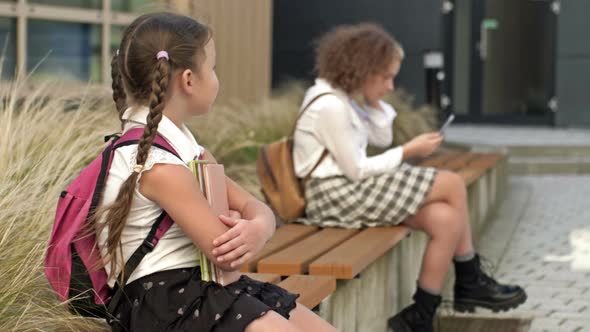 Two Schoolgirls of Different Ages are Sitting on a Bench in the School Yard
