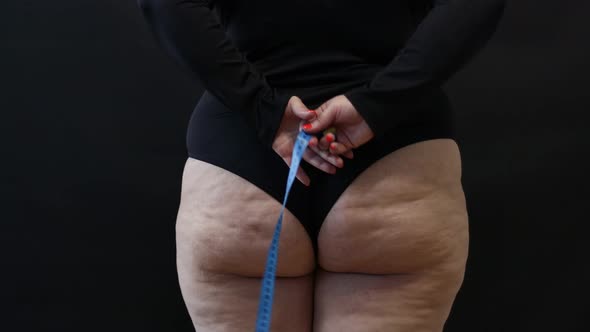 a Fat Woman with Cellulite Measures Herself with a Measuring Tape on a Black Background