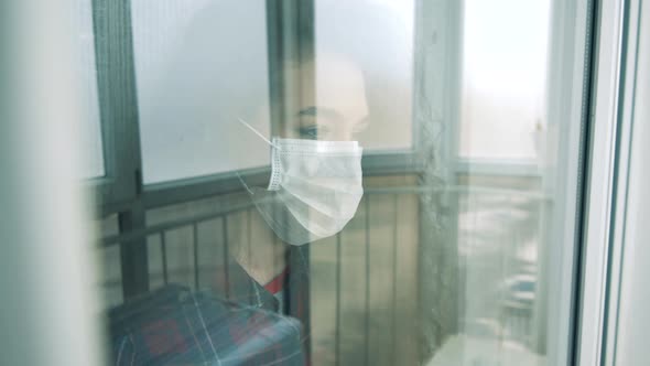 A Woman Wears Medical Mask and Looks Through Window Indoors During Pandemic alt