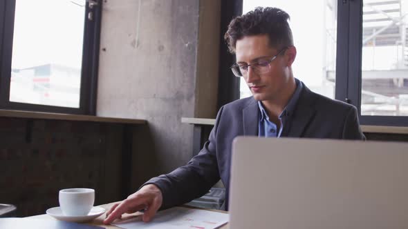 Caucasian businessman using laptop looking at paperwork, sitting at table with coffee in cafe alt