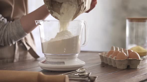 A Girl In A Beige Apron Weighs Flour On A Scale. Flour From A Paper Bag Falls Into A Mechanical Scal alt