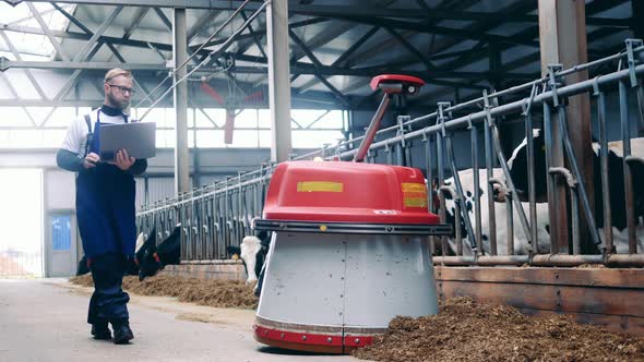Farm Expert with a Laptop is Following an Automated Feed Pusher alt