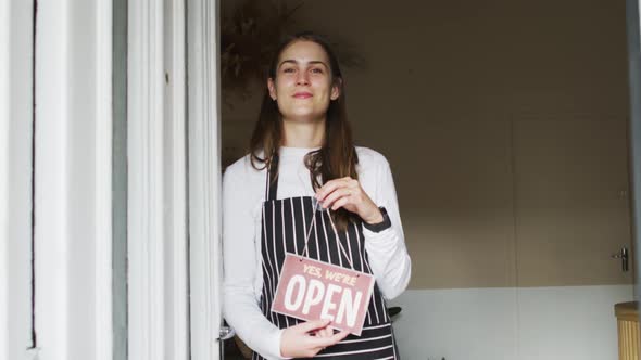 Smiling caucasian waitress standing in door, holding open sign, looking at camera alt