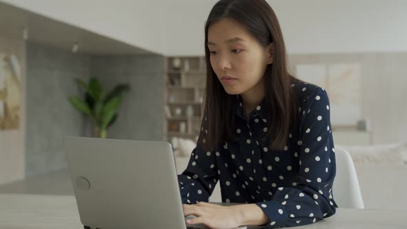 Excited Cheerful Young Asian Woman Sits at a Table with a Laptop Raises Her Hands Up and Celebrates alt