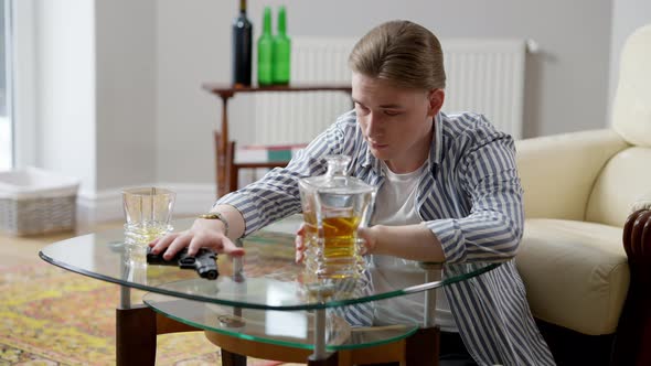Portrait of Desperate Young Man Moving Apart Glass and Decanter with Whiskey Taking Gun in Hand and alt