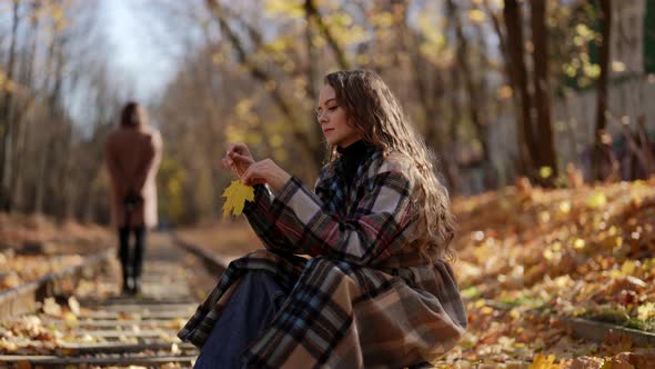a Woman with Curly Hair and a Plaid Coat is Sitting on the Rails with a Yellow Leaf in Hands on a alt
