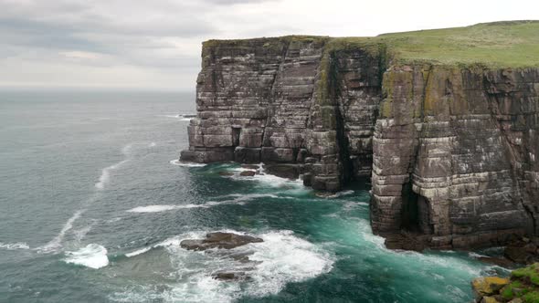 Waves slowly crash over rocks and against a tall and imposing sea cliff in the deep teal coloured oc alt