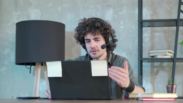 Young Curly Haired Man Using Laptop Computer with Headset to Talking Online alt