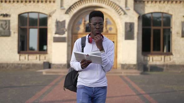 Thoughtful Smart African American Young Man Walking at University Campus Reading Book alt