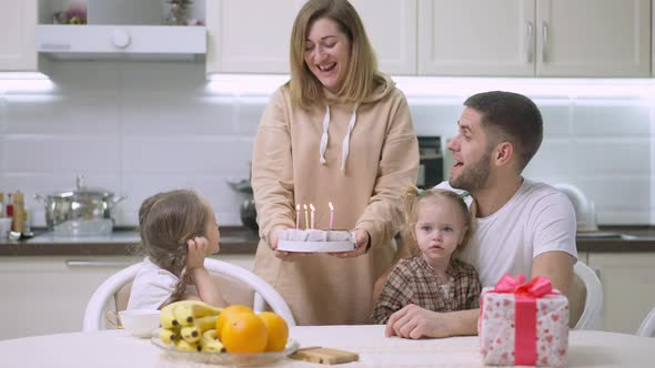 Smiling Mother Holding Birthday Cake As Charming Little Girl Blowing Candles alt
