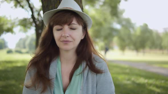 Portrait of a Happy Woman in a Hat Near the Tree in the Park Looking at the Camera