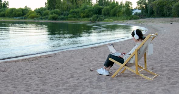 Freelance woman in headphones sitting sideways working on laptop by the lake alt