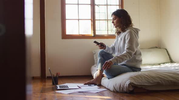 Mixed race woman working at home, sitting on bed using smartphone and laptop in cottage alt