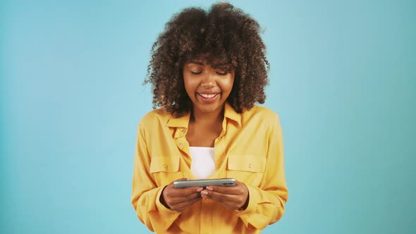Afro American Woman Playing Video Game on Smartphone and Smiling While Posing Against Blue Studio alt