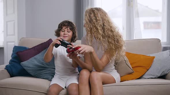 Joyful Mother and Active Nice Little Boy Sitting on Soft Couch and Using Joysticks alt
