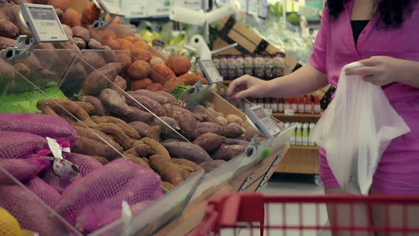 A Young Woman Buys Groceries Picks Up Potatoes in a Bag for Weighing on a Scale in a Supermarket alt