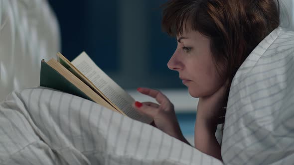 Brunette Woman Covered Herself with a Blanket Lies on the Bed Near the Window and Reads a Book