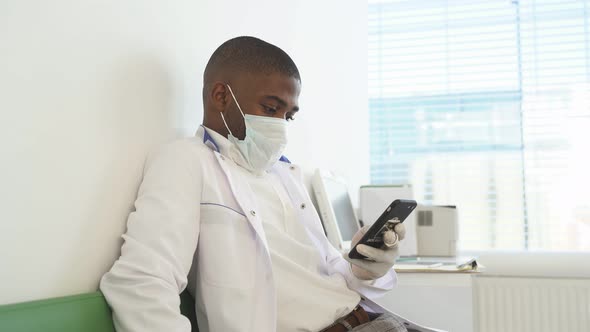 Afro American Male Doctor Sits in Medical Office During Free Time and Working on Smartphone
