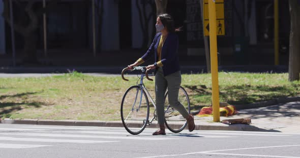African american woman wearing face mask wheeling bicycle crossing road alt