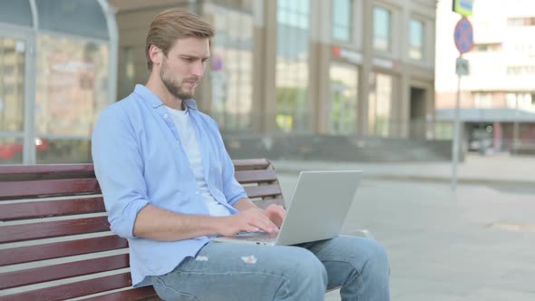 Young Man with Wrist Pain Using Laptop While Sitting on Bench alt