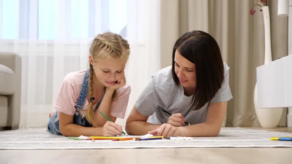 Smiling Young Mom and Cute Preteen Daughter Lying on Warm Floor at Home Drawing in Album Together