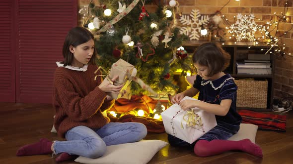 Two Excited Little Girls Shaking Gift Box Under Christmas Tree Sitting on the Floor alt