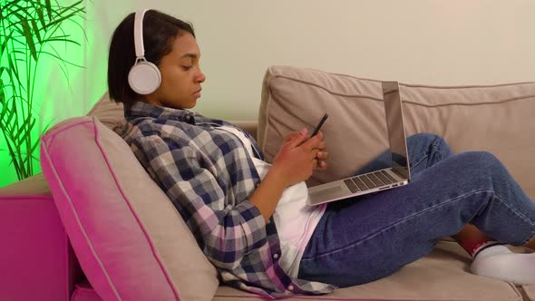 An Woman Lying at Home on the Couch Works Online Using a Laptop and Smartphone