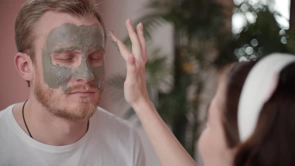 Dating Couple Young Girl Applying Facial Skin Care Mask on Man Boyfriend Face in Cozy Pink Room.