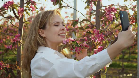 Young Woman in a White Blouse Takes a Selfie Near the Young Trees with Pink Bloom alt