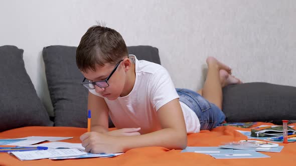 Child Writing with a Pen in a Notebook Lying on an Orange Bedspread in Room alt